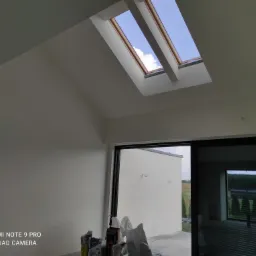 Wnętrze świeżo pomalowanego pokoju z oknami dachowymi i dużym oknem przesuwnym, widoczne worki z zaprawą na podłodze.