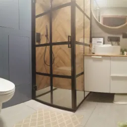 Nowoczesna łazienka z kabiną prysznicową typu walk-in z czarnymi profilami i drewnianym wykończeniem wewnątrz, wisząca toaleta na tle niebieskich płytek, okrągłe lustro nad białą szafką z umywalką.