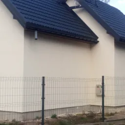 Ściana domu z jasną elewacją, ciemnym dachem i metalowym ogrodzeniem panelowym z widoczną skrzynką gazową.