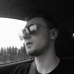 Młody mężczyzna w okularach przeciwsłonecznych typu aviator, siedzący w samochodzie z zapiętym pasem bezpieczeństwa, spoglądający w dal na las za oknem.