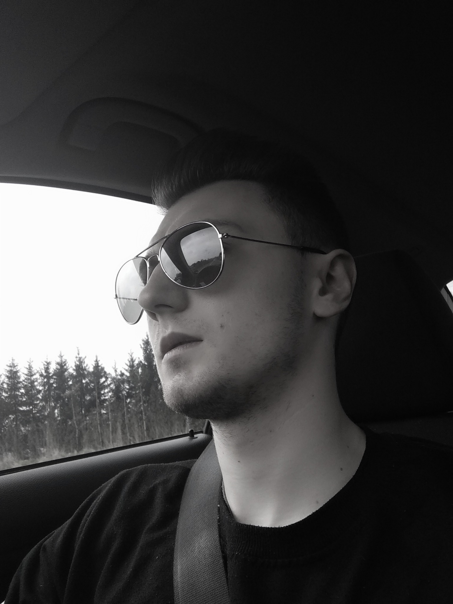 Młody mężczyzna w okularach przeciwsłonecznych typu aviator, siedzący w samochodzie z zapiętym pasem bezpieczeństwa, spoglądający w dal na las za oknem.