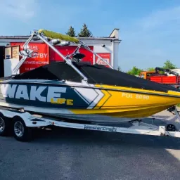 Żółto-szaro-biała łódź motorowa z napisem 'WAKE LIFE' na przyczepie, z czarnym pokrowcem, zaparkowana przed budynkiem z czerwonymi elementami.