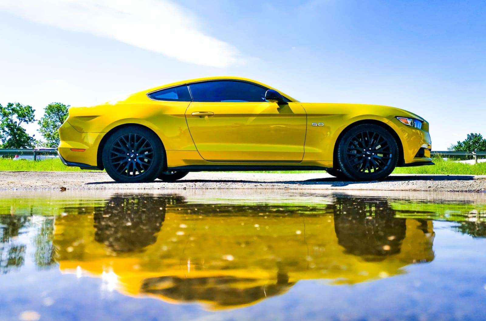 Żółty samochód sportowy Mustang 5.0 z czarnymi felgami, uchwycony w odbiciu kałuży na asfalcie, z błękitnym niebem w tle.