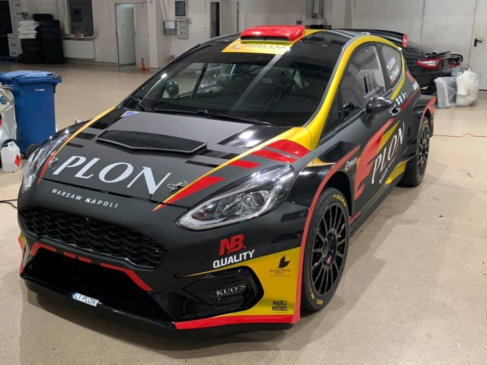 Czarny samochód rajdowy z czerwono-żółtymi akcentami i nazwą 'PLON Warsaw Napoli' na karoserii, sfotografowany w garażu w Warszawie.