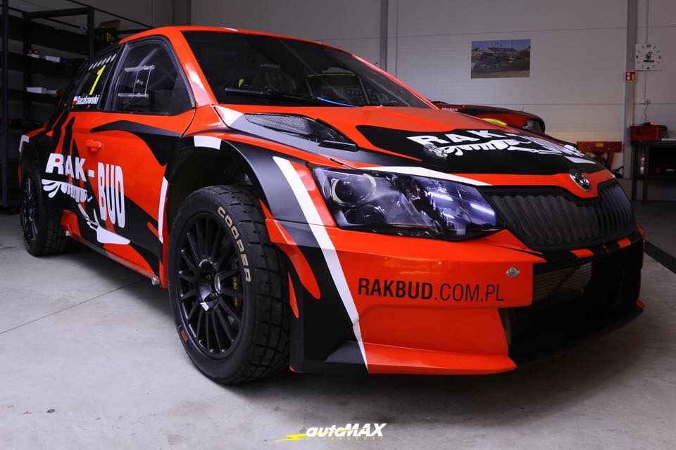 Pomarańczowo-czarny samochód rajdowy Skoda Fabia z naklejkami sponsorskimi RAK-BUD i Cooper Tires, zaparkowany w warsztacie, widok z przodu pod kątem.