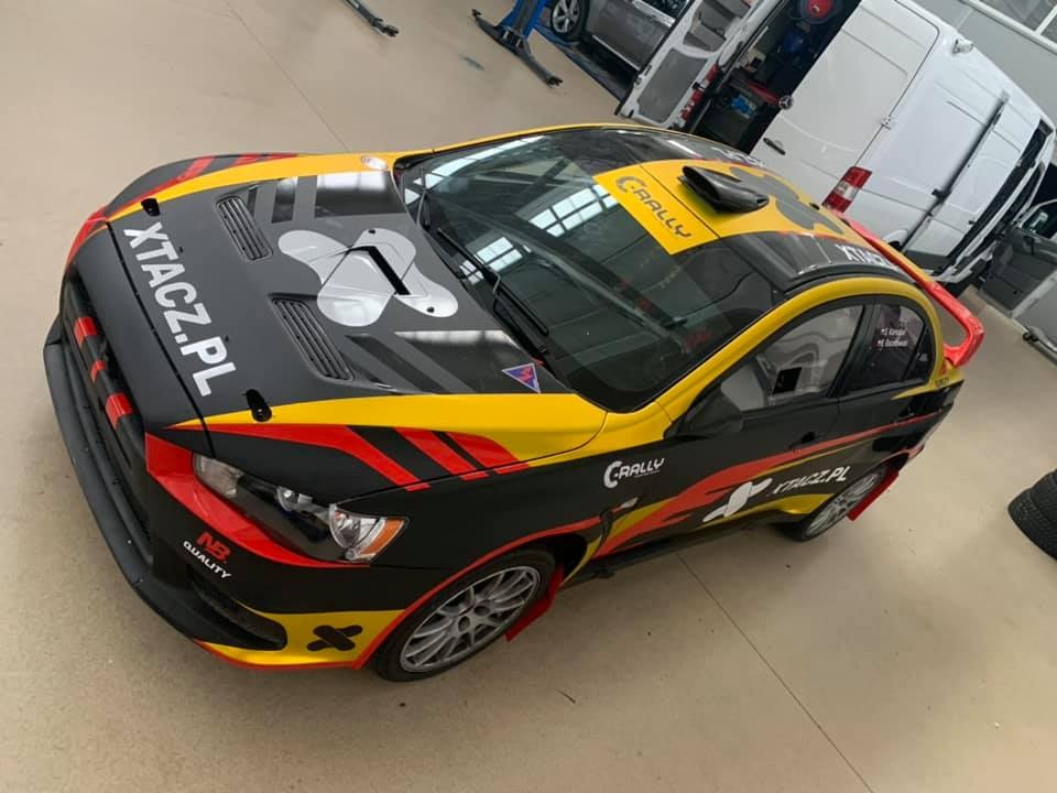 Sportowy samochód Mitsubishi Lancer Evolution w czarno-żółto-czerwonym malowaniu rajdowym z naklejkami i napisami, widoczny z góry w warsztacie samochodowym.