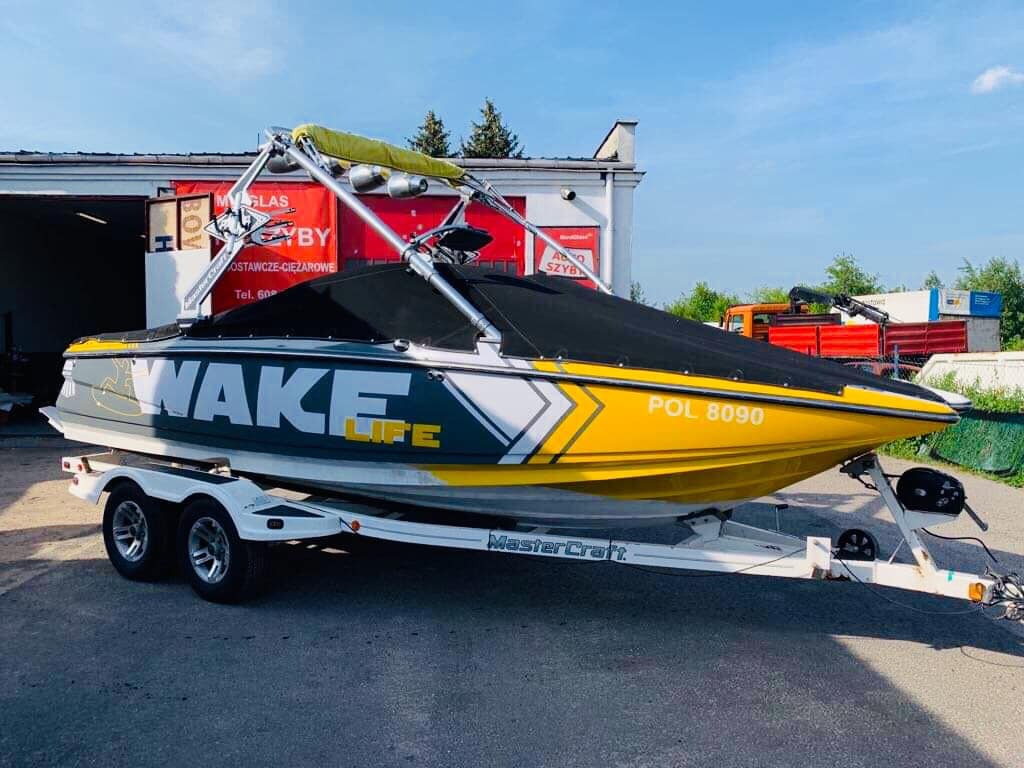 Żółto-szaro-biała łódź motorowa z napisem 'WAKE LIFE' na przyczepie, z czarnym pokrowcem, zaparkowana przed budynkiem z czerwonymi elementami.