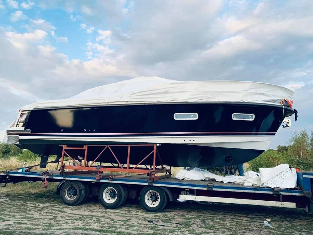Łódź motorowa w kolorze granatowym, częściowo zakryta białą plandeką, transportowana na niskopodwoziowej przyczepie samochodowej, na tle pochmurnego nieba.
