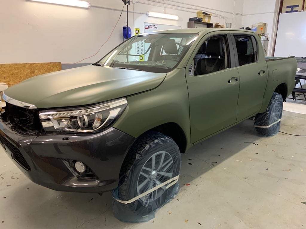 Pickup Toyota Hilux oklejony matową folią w kolorze khaki, z częściowo zdemontowanym przodem i zabezpieczonymi kołami w folii, w warsztacie samochodowym.