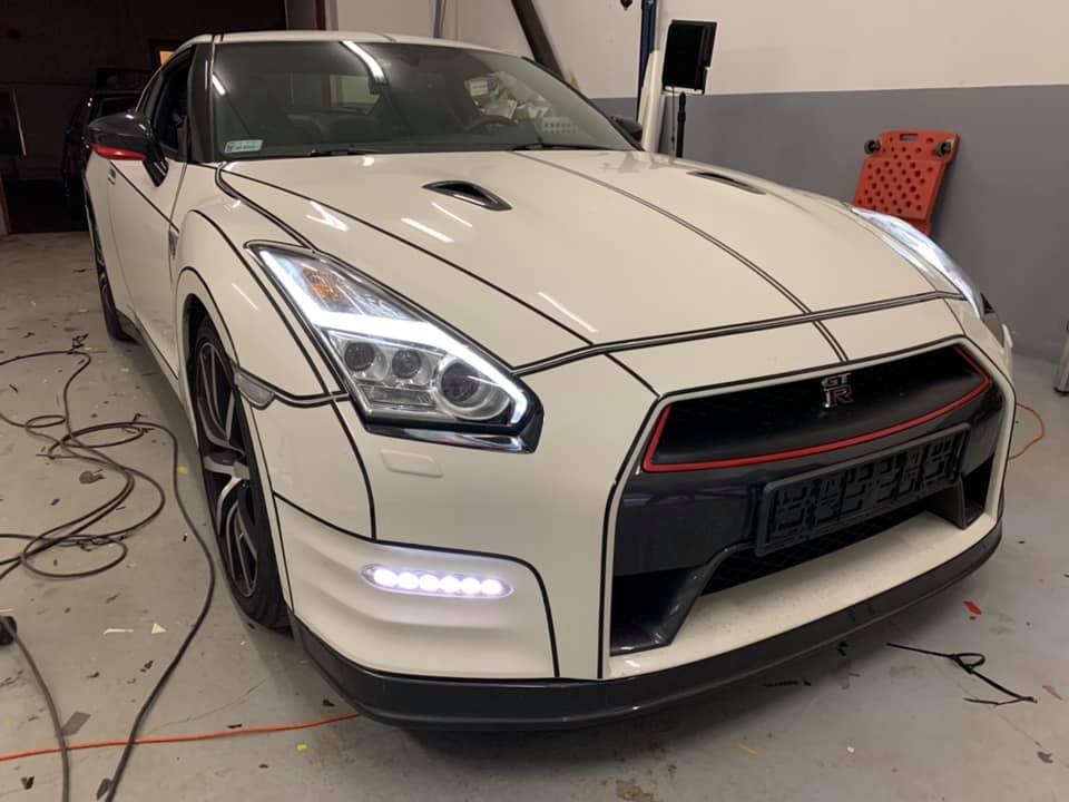 Biały Nissan GT-R z czarnymi konturami w stylu rysunkowym, widoczne światła LED i logo GT-R na grillu, w warsztacie z kablami na podłodze.