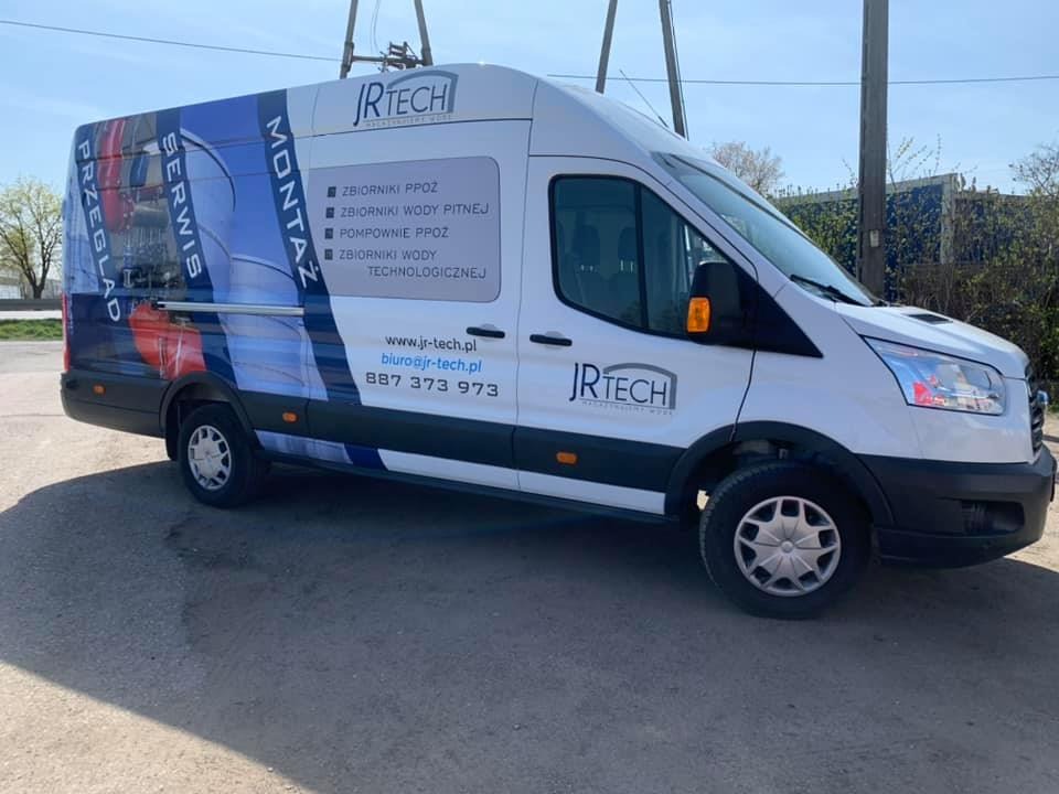 Biały samochód dostawczy z niebiesko-białą grafiką reklamową firmy JRTech, oferującej montaż, serwis i przegląd zbiorników PPOŻ oraz zbiorników wody, zaparkowany na szarej nawierzchni w słoneczny...