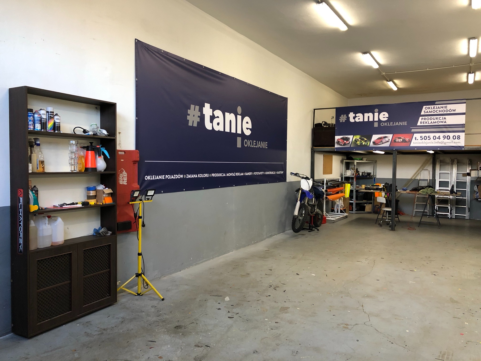 Wnętrze warsztatu z banerem reklamowym '#tanie OKLEJANIE', regałem z narzędziami, motocyklem i stanowiskiem pracy.