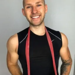 Uśmiechnięty mężczyzna w czarnym tank topie z czerwoną gumą do ćwiczeń wokół szyi, widoczny tatuaż na ramieniu i zegarek na nadgarstku, szarym tło.