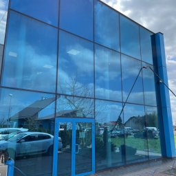 CONSPEKT - Czyszczenie przeszklonej fasady biurowca z użyciem teleskopowej lancy, widoczne refleksy nieba i budynków, na pierwszym planie kostka brukowa i zaparkowane samochody.