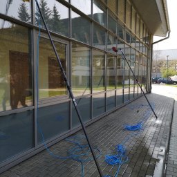 CONSPEKT - Dwa teleskopowe kije z szczotkami do mycia okien na tle przeszklonej fasady budynku biurowego, niebieski wąż doprowadzający wodę leży na mokrej kostce brukowej.