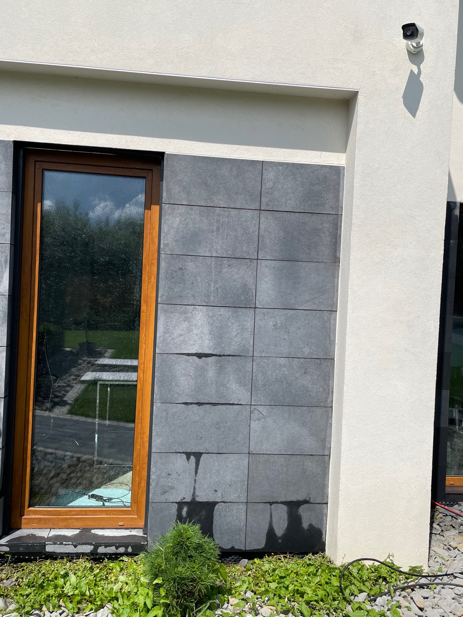 Fragment elewacji budynku z szarymi płytkami i drewnianą ramą okienną; widoczne zacieki na płytkach, roślinność u podstawy ściany i system monitoringu na elewacji.