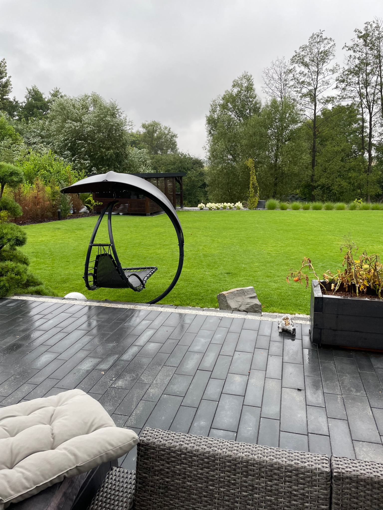 Nowoczesny ogród z huśtawką typu bocianie gniazdo na tle zielonego trawnika i altany, widoczne wilgotne płytki chodnikowe.