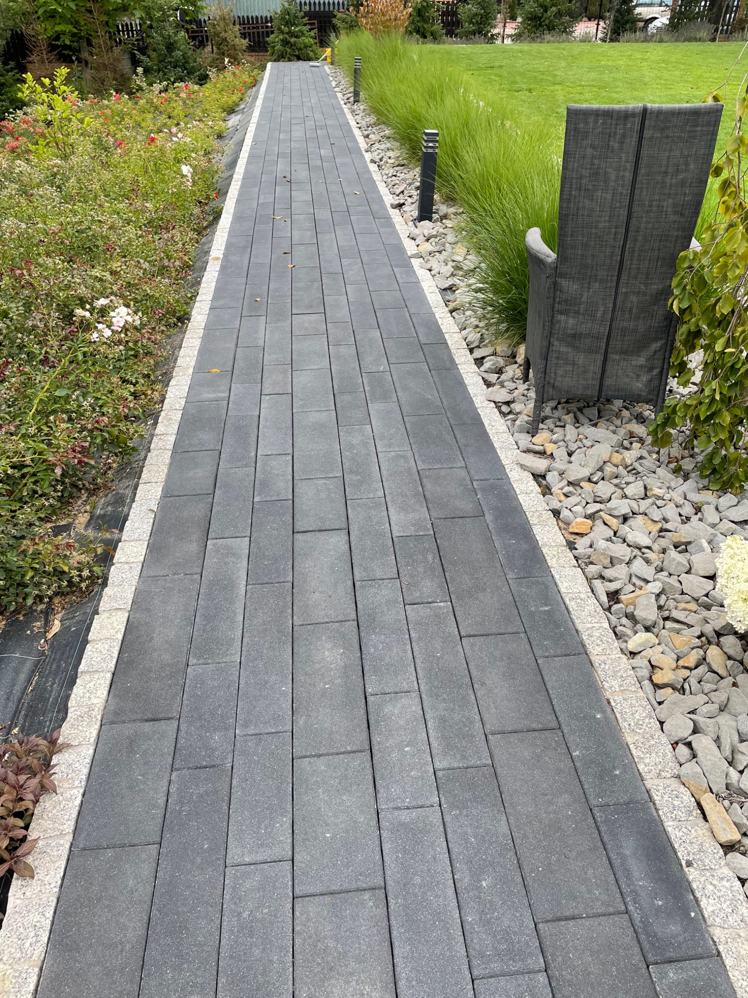 Ścieżka z szarej kostki brukowej obramowana jasnym kamieniem, prowadząca przez zadbany ogród z trawami ozdobnymi i kwitnącymi krzewami, z krzesłem ogrodowym w tle.