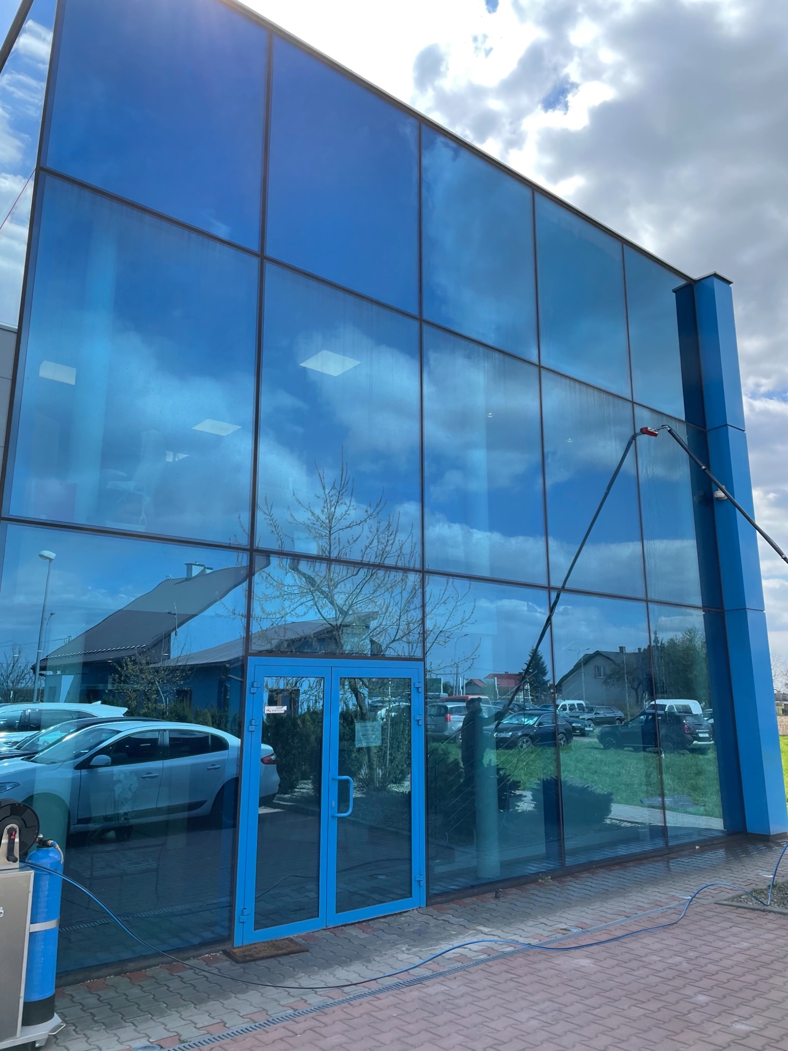 Czyszczenie przeszklonej fasady biurowca z użyciem teleskopowej lancy, widoczne refleksy nieba i budynków, na pierwszym planie kostka brukowa i zaparkowane samochody.