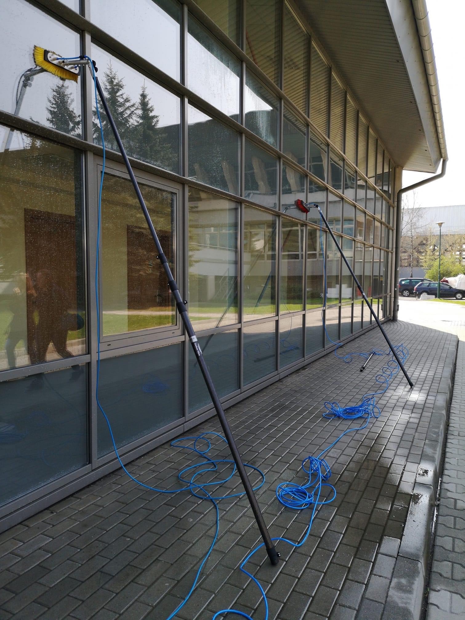 Dwa teleskopowe kije z szczotkami do mycia okien na tle przeszklonej fasady budynku biurowego, niebieski wąż doprowadzający wodę leży na mokrej kostce brukowej.