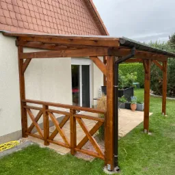 Brązowa, drewniana konstrukcja z zadaszeniem, przylegająca do ściany domu, z dekoracyjną balustradą i widoczną rynną odprowadzającą wodę, otoczona zielonym trawnikiem.