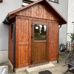 Drewniana zabudowa wejścia do domu z przeszklonymi drzwiami, widoczne elementy orynnowania, betonowe schodki i czarna wycieraczka przed wejściem.