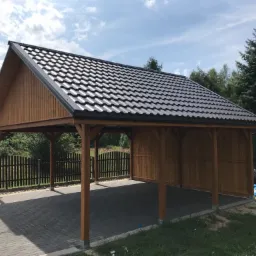 Drewniana konstrukcja z zadaszeniem z blachy trapezowej, służąca jako wiata garażowa, na posesji z kostką brukową i ogrodzeniem.