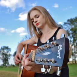 Martyna Święcicka Fotografia - Młoda kobieta z blond włosami gra na gitarze akustycznej marki Cort na tle błękitnego nieba z chmurami i zielonego pola.