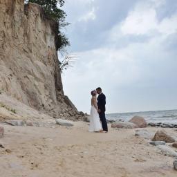 Martyna Święcicka Fotografia - Para młoda w objęciach na piaszczystej plaży z klifem w tle, morze w oddali pod pochmurnym niebem.