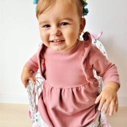 Martyna Święcicka Fotografia - Uśmiechnięta dziewczynka w różowej sukience i błękitnym wianku siedzi na białym, plecionym krzesełku z kwiatowym motywem.