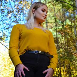 Martyna Święcicka Fotografia - Portret kobiety w żółtej bluzce i czarnych jeansach z dziurami na kolanach, pozującej na tle jesiennego lasu z niebem w tle, z widocznymi drzewami i liśćmi.