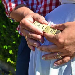 Przyszli rodzice trzymają na brzuchu kobiety klocki z imieniem 'Kalina', ona ma pierścionek na palcu i ozdobiony paznokieć, on ma koszulę w czerwoną kratę.