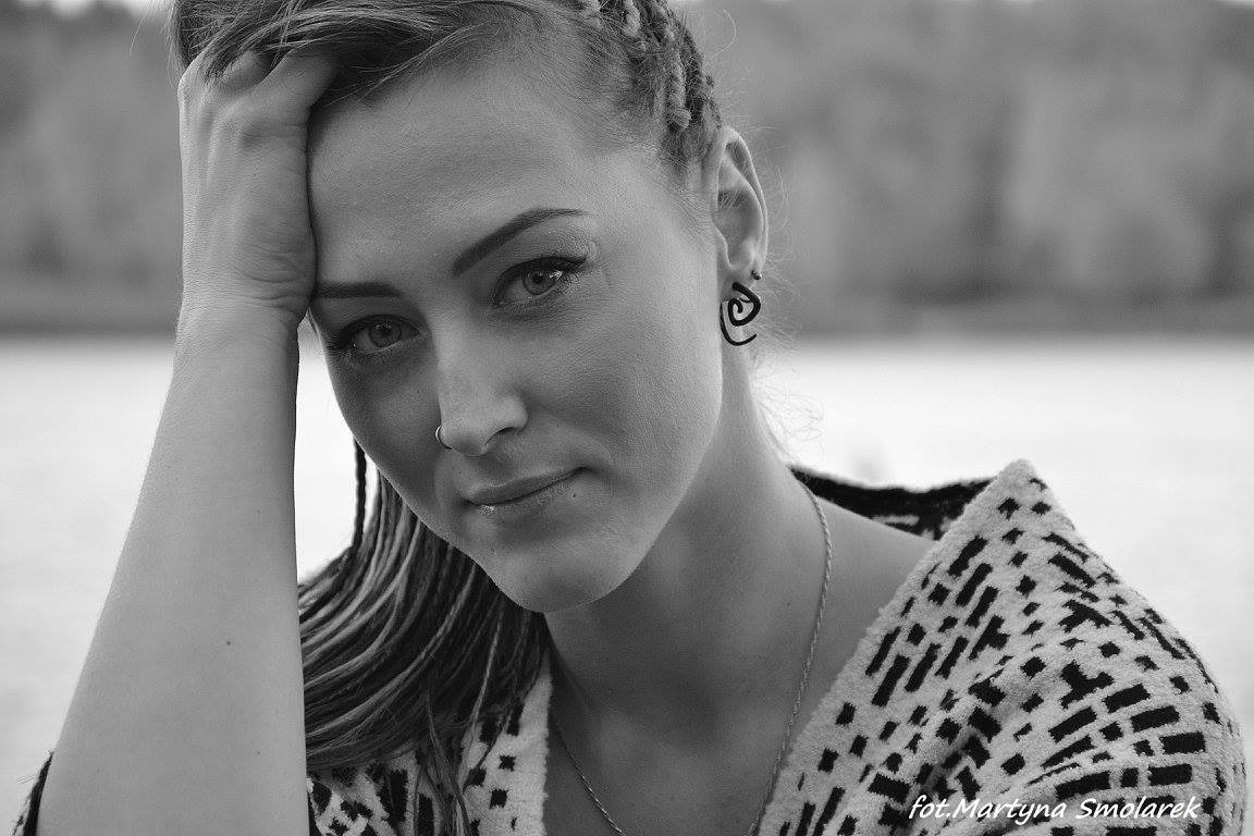 Portret kobiety w czarno-białej tonacji, z ręką opartą o głowę, noszącej kolczyk w nosie i geometryczne kolczyki, na rozmytym tle.