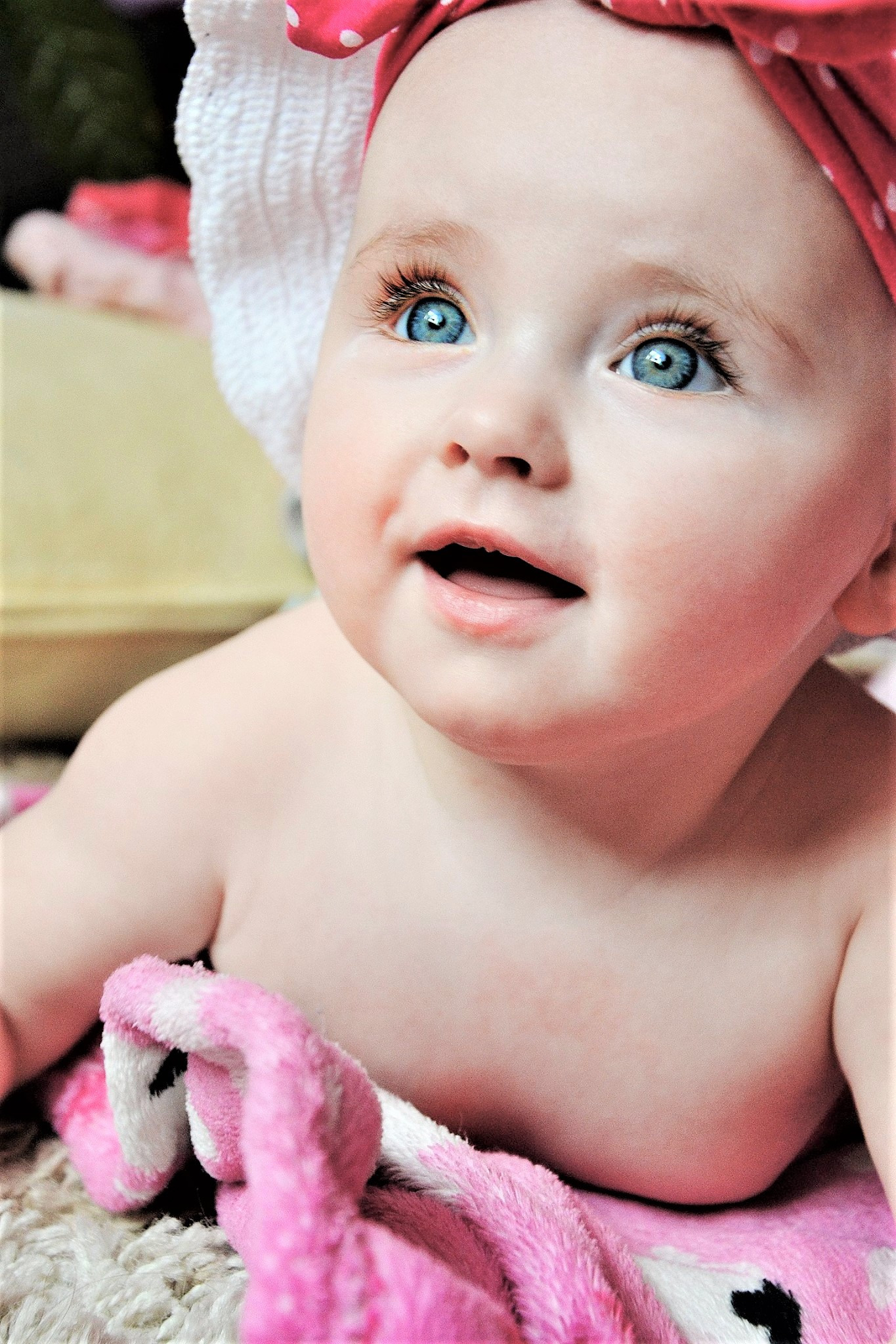 Niemowlę z dużymi niebieskimi oczami, ubrane w czerwoną opaskę w białe kropki, leży na brzuchu na białym dywanie z różowym kocykiem.
