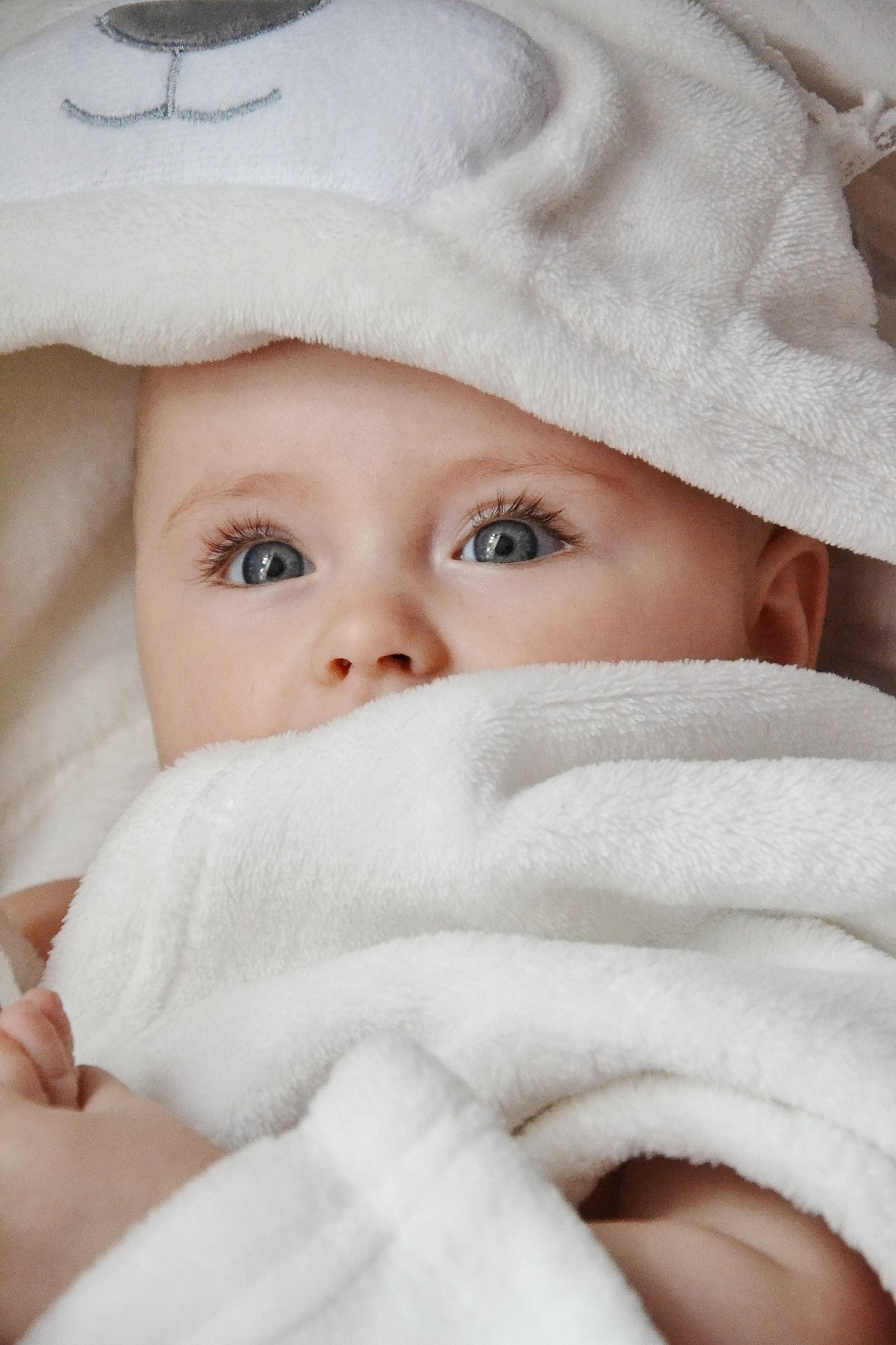 Niemowlę z dużymi, niebieskimi oczami, owinięte w biały, puszysty ręcznik z kapturem w kształcie misia.