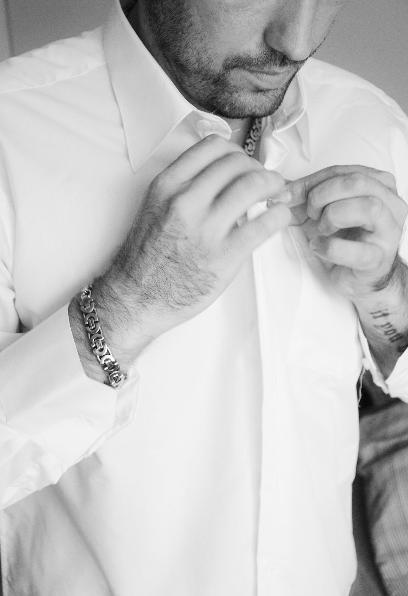 Czarno-białe zdjęcie mężczyzny zapinającego białą koszulę, widoczny srebrny łańcuszek na nadgarstku i tatuaż.
