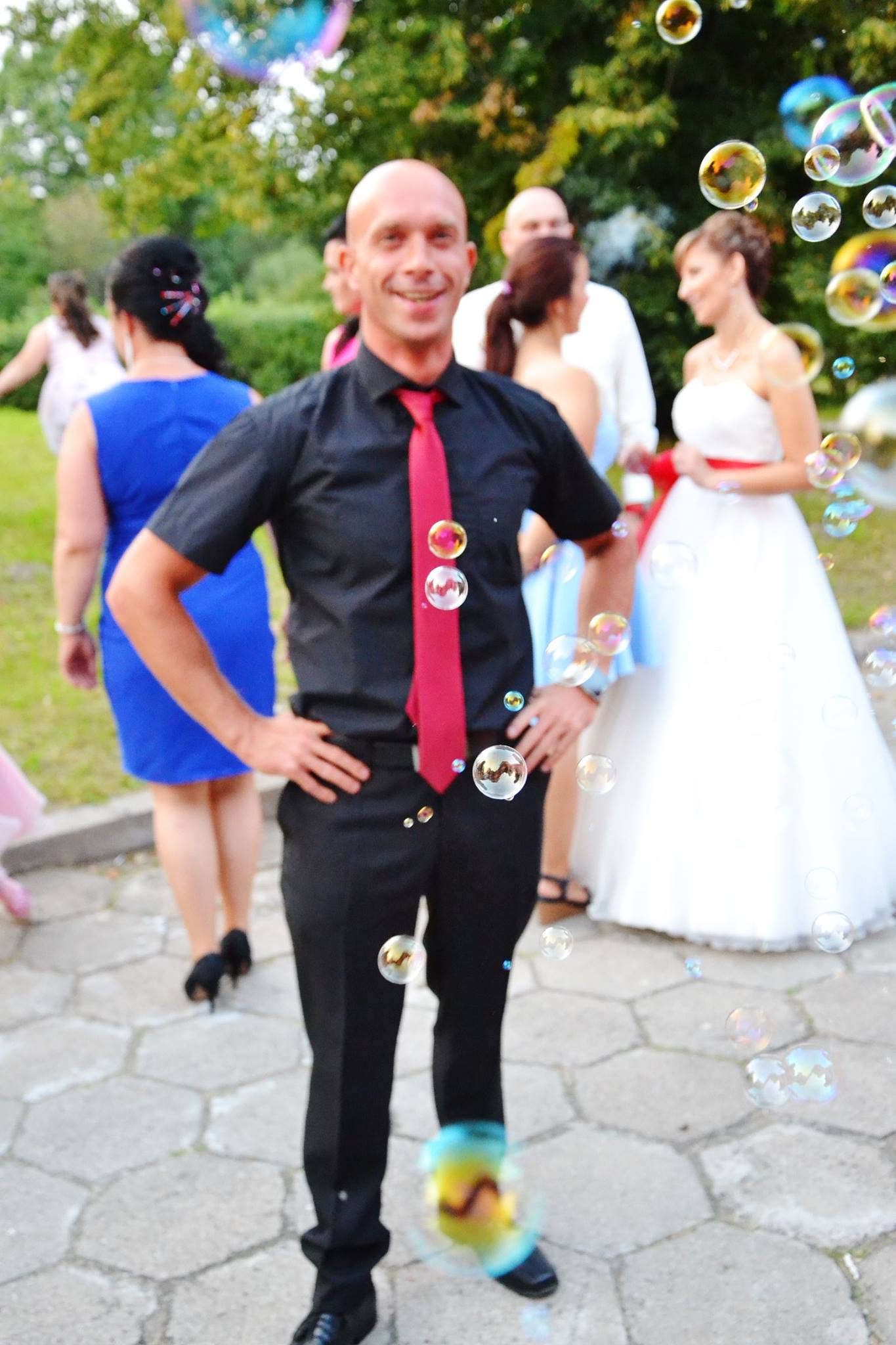 Uśmiechnięty mężczyzna w czarnej koszuli i czerwonym krawacie, otoczony bańkami mydlanymi, w tle para młoda i goście weselni.