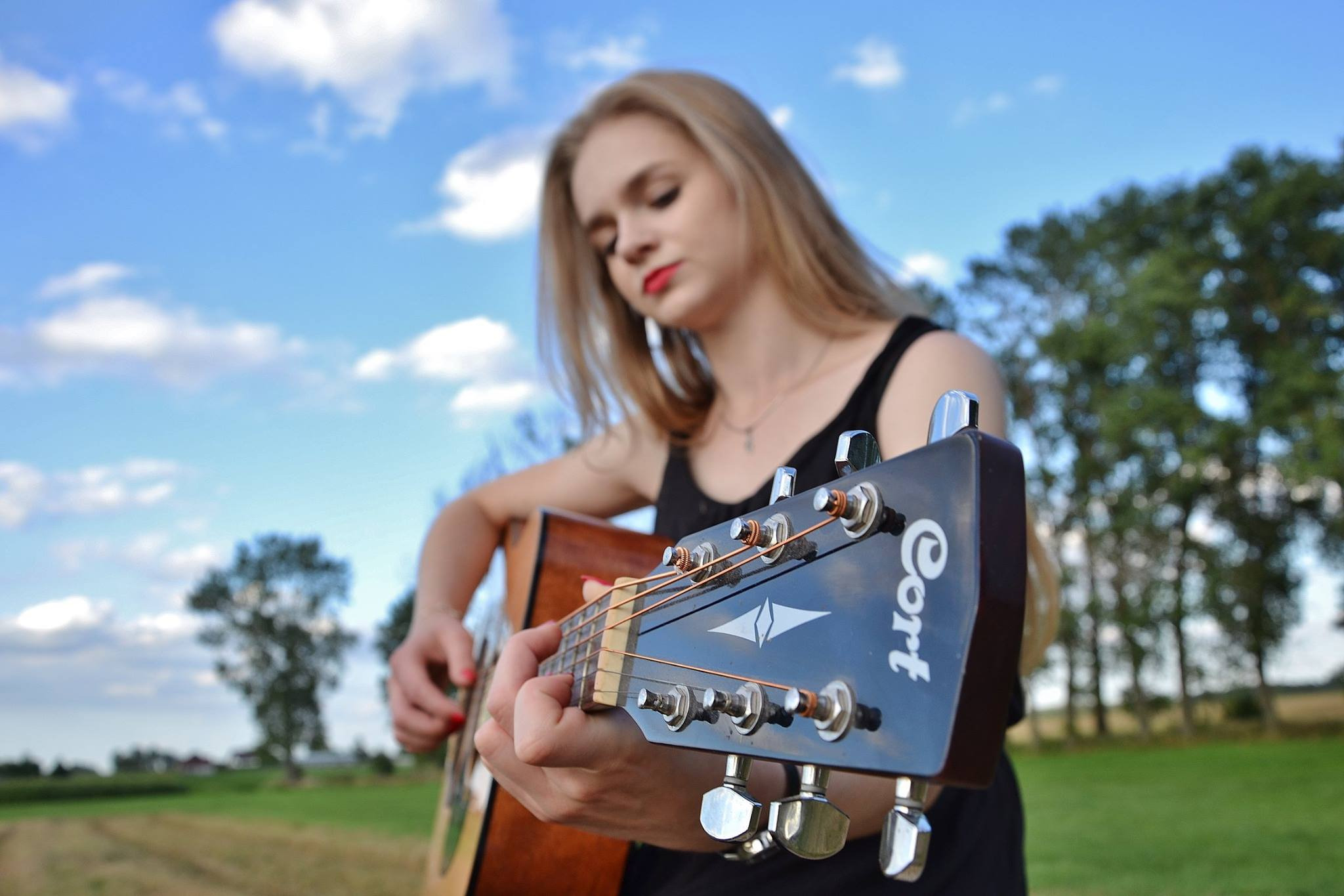 Młoda kobieta z blond włosami gra na gitarze akustycznej marki Cort na tle błękitnego nieba z chmurami i zielonego pola.