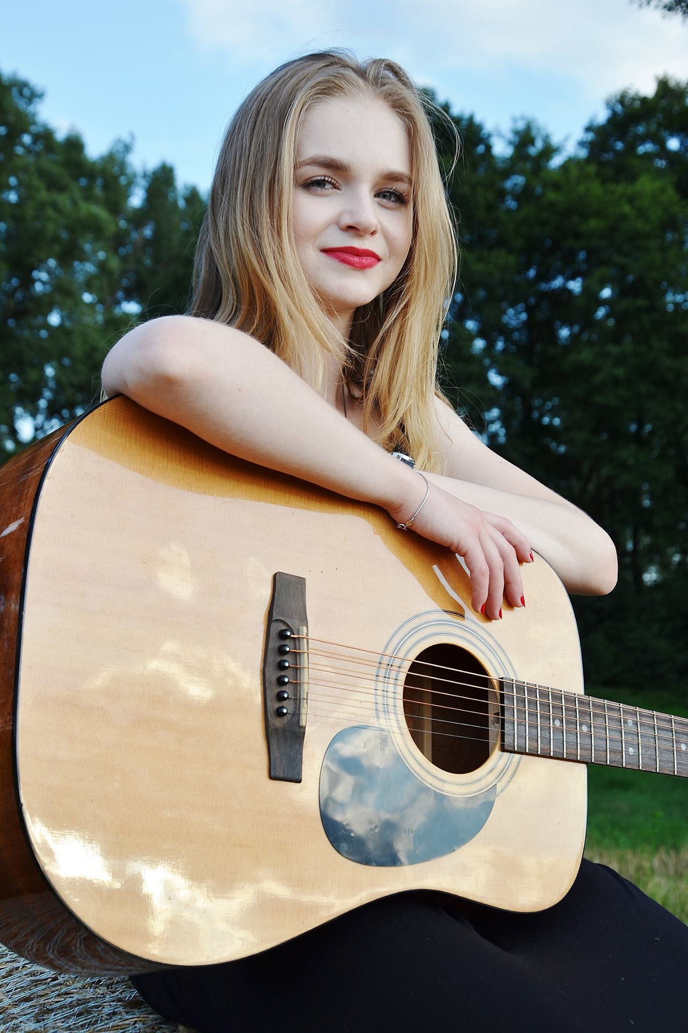 Portret młodej kobiety z blond włosami i czerwonymi ustami, opartej o akustyczną gitarę na tle zieleni.