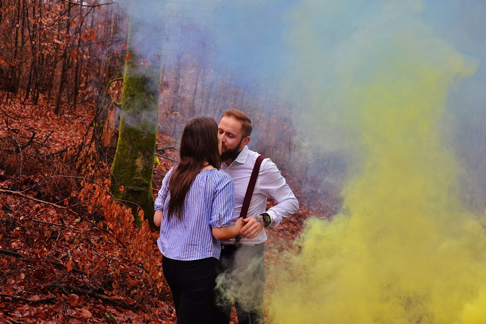 Para całująca się w lesie, otoczona kolorowym dymem, drzewa w tle z brązowymi liśćmi, mężczyzna w białej koszuli z szelkami, kobieta w bluzce w paski.
