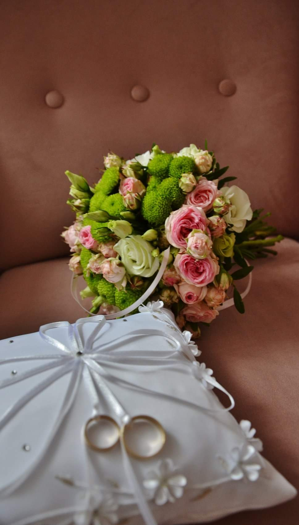 Bukiet ślubny z różowych i zielonych kwiatów oraz obrączki na białej poduszce z kokardami, na tle brązowego fotela z guzikami.