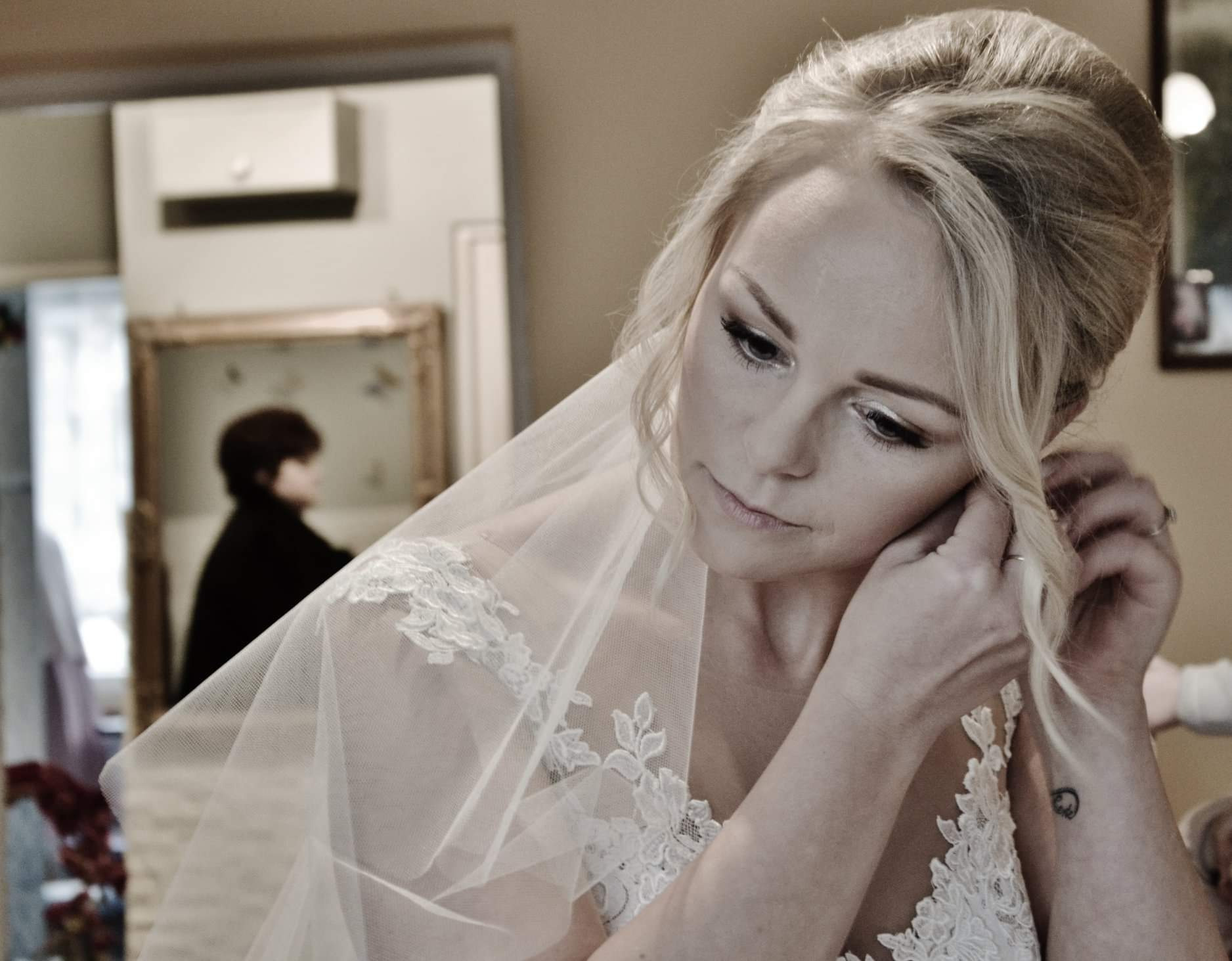 Panna młoda w białej sukni z koronkowymi zdobieniami i welonem, poprawia kolczyk, widoczny tatuaż na nadgarstku, w tle lustro z odbiciem osoby.