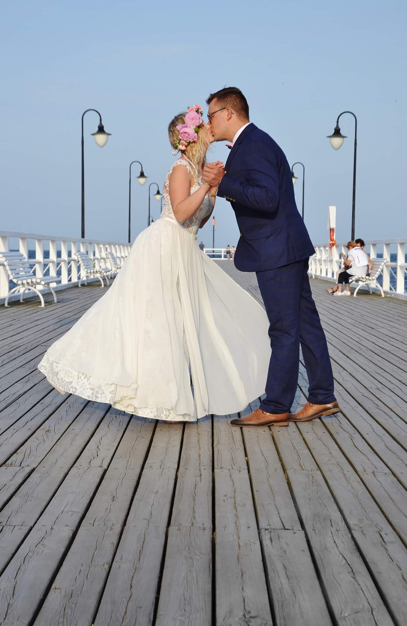 Para młoda całuje się na drewnianym molo; panna młoda w białej sukni z koronkowym wykończeniem i wiankiem z różowych kwiatów, pan młody w granatowym garniturze w kratę i brązowych butach.