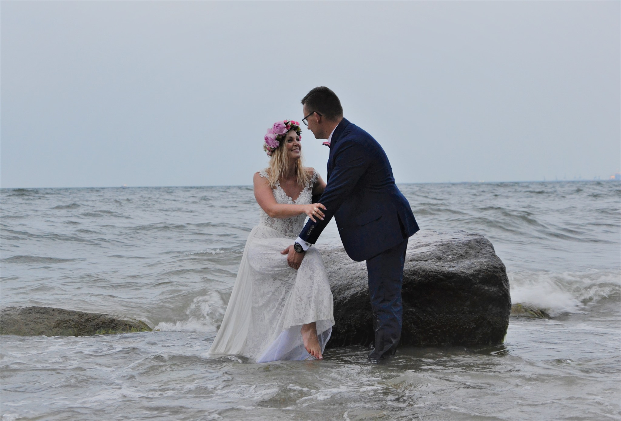 Para młoda na kamieniu w morzu: panna młoda w koronkowej sukni i wianku z różowych kwiatów, pan młody w granatowym garniturze z muszką, oboje patrzą na siebie z uśmiechem, w tle lekko zamglone morze.