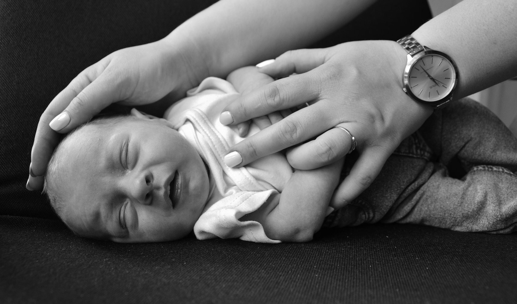 Czarno-białe zdjęcie śpiącego noworodka, którego głowę i ciało otulają dłonie kobiety z zegarkiem na ręce i pierścionkiem na palcu.