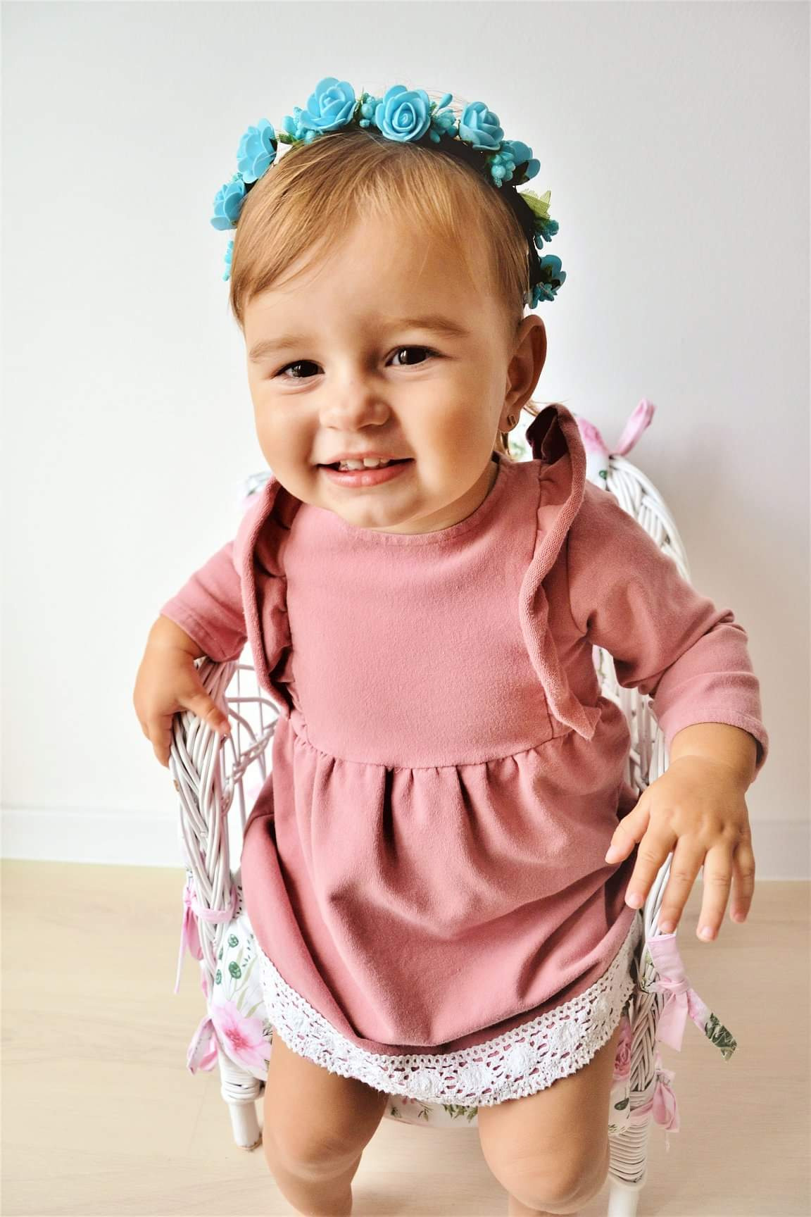 Uśmiechnięta dziewczynka w różowej sukience i błękitnym wianku siedzi na białym, plecionym krzesełku z kwiatowym motywem.