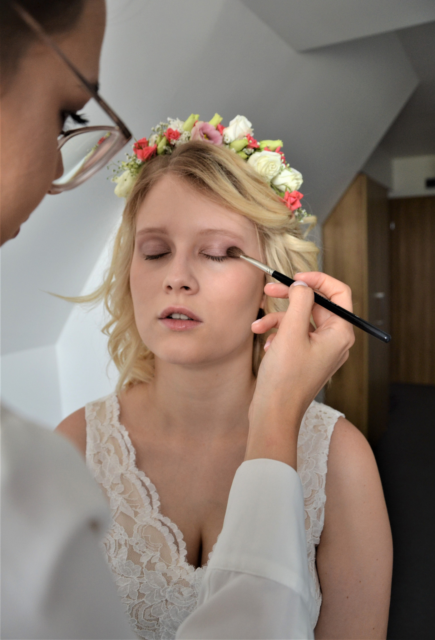 Panna młoda z wiankiem na głowie podczas wykonywania makijażu oka. Makijażystka używa pędzla. Delikatny makijaż ślubny, koronkowy dekolt sukni.