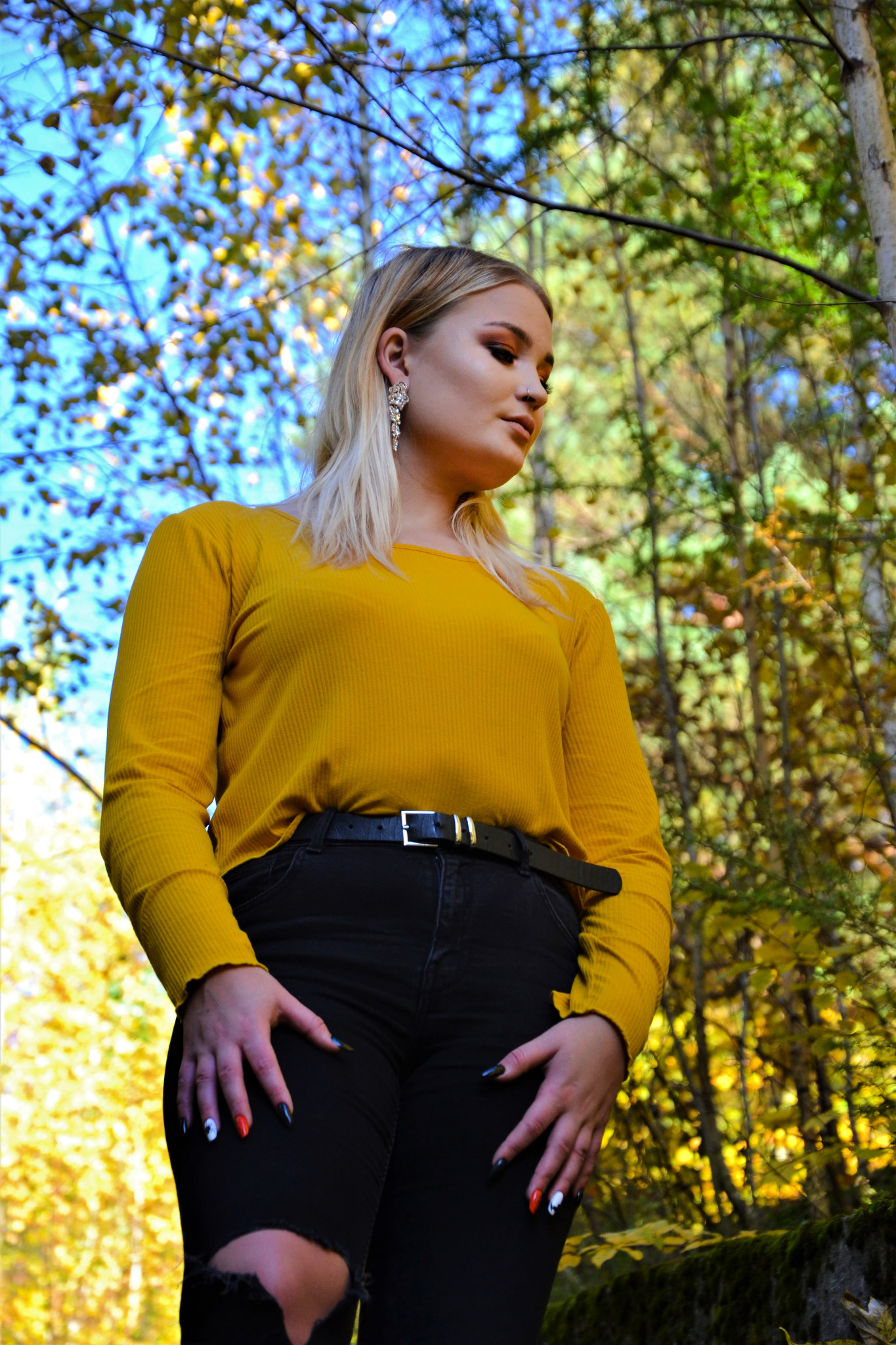 Portret kobiety w żółtej bluzce i czarnych jeansach z dziurami na kolanach, pozującej na tle jesiennego lasu z niebem w tle, z widocznymi drzewami i liśćmi.