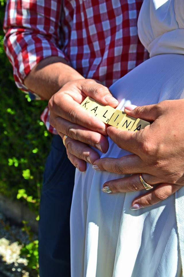 Przyszli rodzice trzymają na brzuchu kobiety klocki z imieniem 'Kalina', ona ma pierścionek na palcu i ozdobiony paznokieć, on ma koszulę w czerwoną kratę.