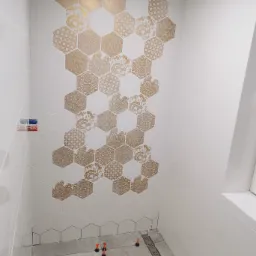 Ściana w łazience wyłożona białymi heksagonalnymi płytkami z dekoracyjnymi, złotymi wzorami, widoczny odpływ liniowy w podłodze i system poziomowania płytek.